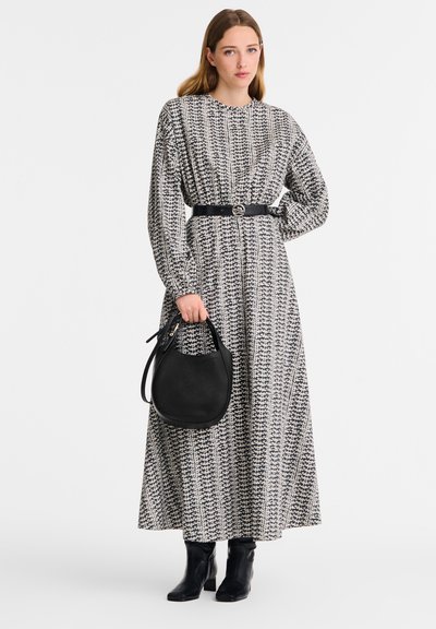 Langärmliges gemustertes Kleid in Schwarz und Weiß, tailliert mit einem schwarzen Gürtel. Das Model hält eine schwarze strukturierte Handtasche und trägt schwarze Knöchelstiefel.
