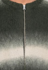 Close-up de uma peça de vestuário de malha felpuda com tons degradê de verde-escuro para cinza-claro e um fecho de zíper metálico prateado.