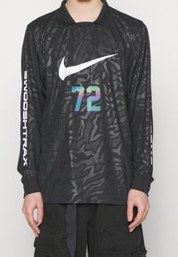 T-shirt noir à manches longues Nike avec un swoosh blanc, le numéro 72 irisé sur le devant, et le texte "SWOOSHTRAX" en blanc sur les manches, porté avec un pantalon noir.