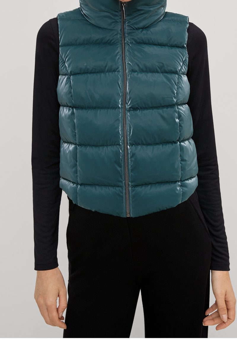 Doudoune sans manches couleur sarcelle avec col montant, design matelassé, fermeture éclair à l'avant et finition brillante ; portée par-dessus un haut noir à manches longues.