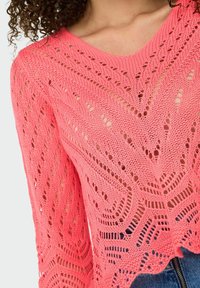 Pull en tricot crochet de couleur corail vif. Il présente un col en V, des manches longues, et une texture à motifs avec des sections ajourées et un ourlet festonné.
