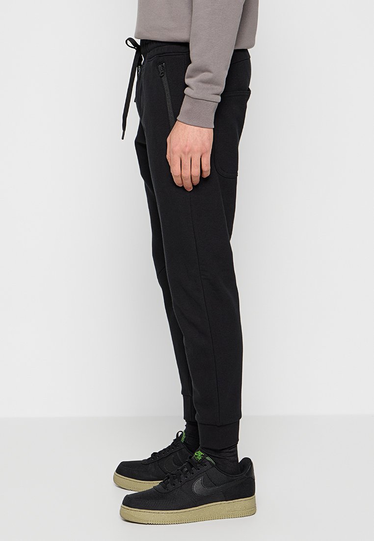 Pantaloni sportivi neri con un design affusolato, vita elastica con cordino e tasche laterali con zip, abbinati a sneakers nere Nike.