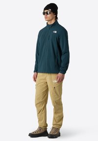 Teal zip-up jacket, beige cargo pants, and brown hiking boots. The outfit features branding and a snug beanie, designed for outdoor activities.