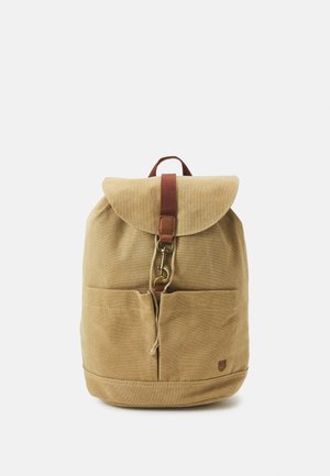 Sac à dos en toile beige avec un rabat arrondi, une sangle en cuir marron et deux poches avant. Présente un design minimaliste avec un détail de logo.
