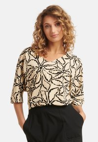 Crème blouse met een zwart bladenpatroon, V-hals en driekwart mouwen, gecombineerd met zwarte broek. Gladde stoftextuur.