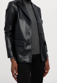 Svart konstläderblazer med en strukturerad design, slagkragar, två framknappar och en ficka på vänster sida.