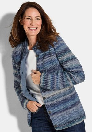 Veste en bleu texturé avec des rayures horizontales, dotée d'un front boutonné, d'un col rond et d'un tissu doux. Portée par-dessus un haut blanc.