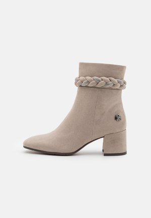 Bottine beige en daim avec petit talon bloc, bride tressée ornée de strass autour de la cheville, et petit emblème floral métallique près du talon.