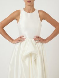 Γυναίκα που φοράει άσπρο σατέν φόρεμα χωρίς μανίκια με στενό κορσέ και φούστα με πτυχώσεις, χέρια τοποθετημένα στους γοφούς σε ουδέτερο φόντο.