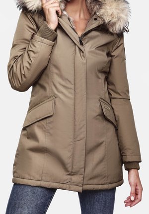 Femme portant un parka d'hiver beige avec capuche bordée de fourrure, poches à rabat à l'avant et poignets côtelés, tenant la capuche près de son visage.