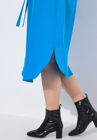 Kirkas sininen mekko, jossa on korkea halkio ja hulmuava muotoilu. Mustat nilkkurit, joissa on kiiltävä krokotiili-tekstuuri ja paksut korot.