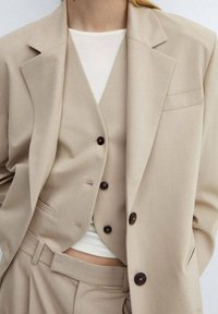 Beżowy szyty na miarę garnitur z jednorzędową marynarką, dopasowaną kamizelką z guzikami oraz białą bluzką pod spodem. Gładka struktura tkaniny.