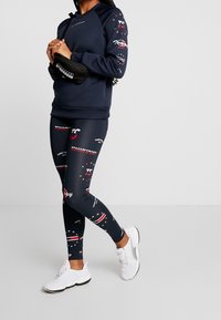 Sudadera navy con mangas estampadas y acentos negros, combinada con leggins ajustados que presentan un estampado de logo en rojo y blanco; zapatillas blancas.