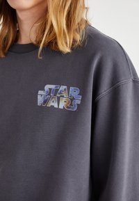 Sweatshirt grigio scuro con scollo rotondo, caratterizzato da una grafica "STAR WARS" blu e multicolore stampata nella parte sinistra del petto.