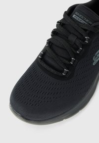 Gros plan sur une chaussure de course Skechers Flex noire avec une tige en mesh respirant, des lacets noirs et une semelle grise sur fond blanc.
