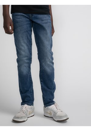 Jeans di denim blu a taglio slim, con una leggera scoloritura e dettagli a effetto whiskering. Abbinati a sneakers bianche con accenti grigi e dorati.