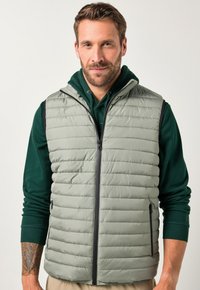 Graue, gepolsterte Weste mit vertikal gestepptem Muster, Frontreißverschluss und zwei Seitentaschen. Über einem dunkelgrünen Hoodie getragen. Glatte Stoffoberfläche.