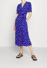 Blue wrap dress with puff sleeves and a belt, featuring a pattern of small colorful circles and a ruffled hem. Worn with black strappy sandals.