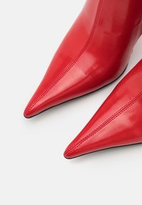 Red patent leather high-heeled shoes with pointed toes and contrasting stitching. Smooth texture with a sleek design and black soles.