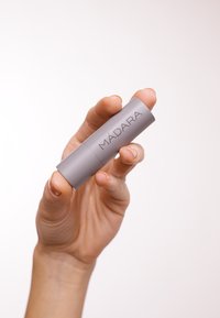 Balsam do ust Mádara w eleganckiej, cylindrycznej, szarej tubce z matowym wykończeniem, z nadrukowanym logo. Trzymany między palcami.