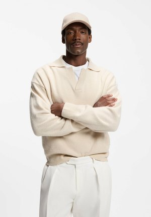 Mies, jolla on päällään kerma-valkoinen villapaita, valkoiset housut ja beige lippalakki, seisoo käsivarret ristissä valkoista taustaa vasten.