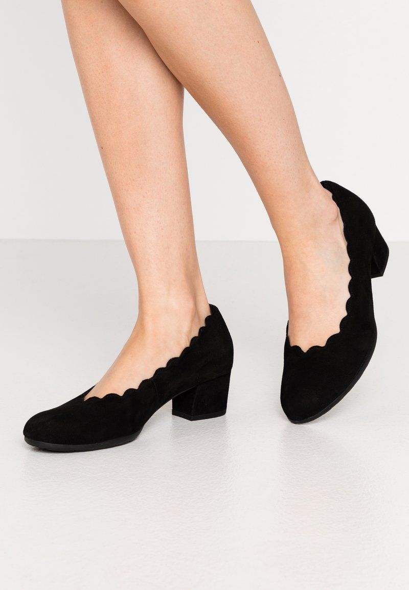 Femme portant des chaussures à talons carrés en daim noir avec des bords festonnés, debout sur un sol gris clair contre un fond blanc uni.