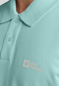 Lichtgroene polo met textuurstof, drie knopen bij de kraag, en een wit logo op de borst met een pootafdruk.