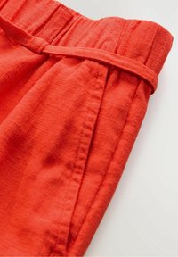 Gros plan sur la ceinture d’un pantalon en lin rouge avec une boucle de ceinture en tissu assorti et une couture visible de la poche latérale.