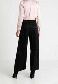 Schwarze weite Hosen mit einem strukturierten Zickzackmuster, kombiniert mit einer hellrosa, langärmligen, glänzenden Bluse und schwarzen High-Heels.