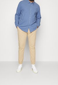 Niebieska koszula na długi rękaw z guzikiem, beżowe spodnie zwężane z elastycznymi mankietami oraz białe trampki. Prosty design, bawełniany materiał.