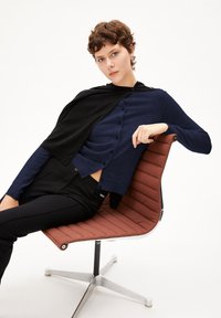 Navyblauer Langarmoberteil mit Knopfdetails, unter einer schwarzen Strickjacke getragen, kombiniert mit schwarzen Hosen, sitzend auf einem braunen, gerippten Stuhl.
