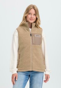 Femme portant un gilet en polaire beige avec une poche poitrine zippée étiquetée "Conçu avec dévouement" sur un sweat-shirt blanc et un jean bleu.