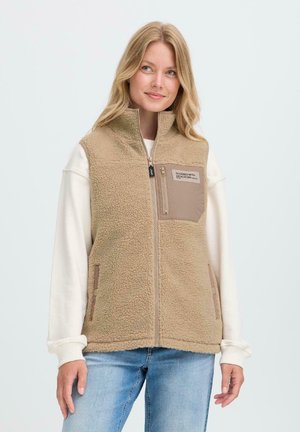 Kvinne som har på seg en beige fleecevest med en glidelås-lomme på brystet merket "Designet med dedikasjon" over en hvit genser og blå jeans.