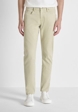 Man die beige slim-fit jeans en witte sneakers draagt, staand tegen een effen witte achtergrond.