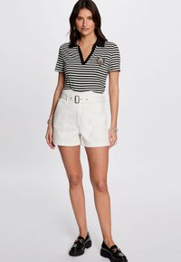 Zwart-wit gestreepte polo met v-hals en badge-detail, gecombineerd met witte shorts met riem. Schoenen zijn zwarte loafers met dikke zolen.