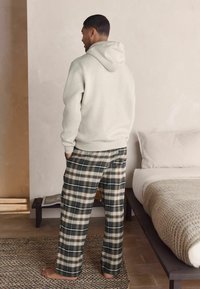 Hanorac gri deschis cu glugă și șnur, asortat cu pantaloni de pijama în carouri verde și bej. Decorul include un pat și un covor țesut.