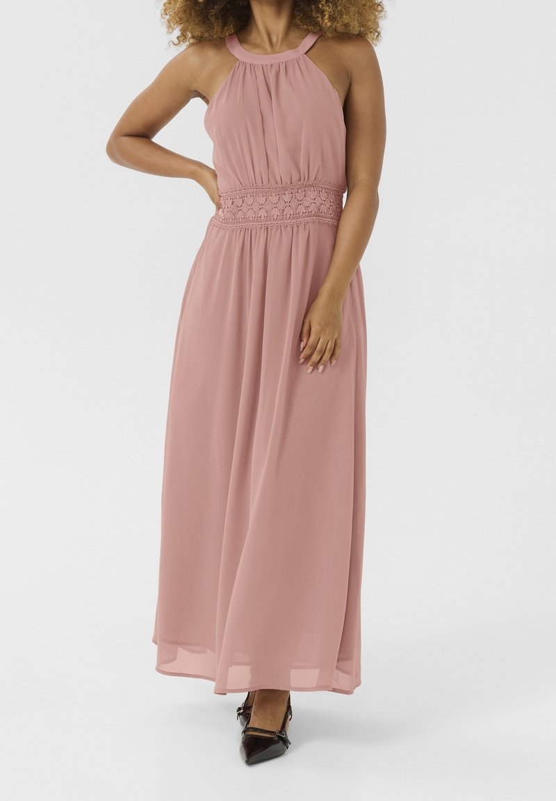 Femme portant une robe longue rose sans manches avec une ceinture en dentelle et des chaussures noires pointues, debout sur un fond uni.
