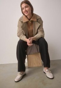 Beige trench coat egy bézs pulóver felett, fekete nadrággal és világos színű mokaszinnal, arany kiegészítéssel. Egyszerű háttér.