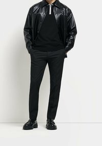 Mężczyzna w czarnej skórzanej kurtce, czarnym swetrze na zamek, czarnych spodniach i czarnych skórzanych mokasynach stoi przy białej ścianie.