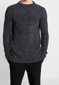 Tmavě šedý pletený svetr s kulatým výstřihem, žebrovanými manžetami a lemem. Tkanina má skvrnitý vzor s bílými akcenty.