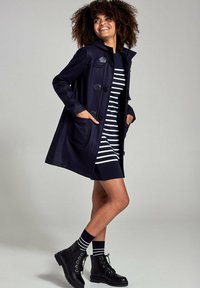 Marineblauwe wollen mengeling jas met toggle sluitingen, gedragen over een zwart-wit gestreepte jurk. Zwarte veterschoenen tot de enkel en gestreepte sokken zichtbaar.