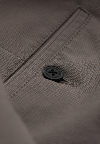 Tissu marron texturé avec un bouton noir à quatre trous et de subtils détails de couture autour d'une petite ouverture de poche.