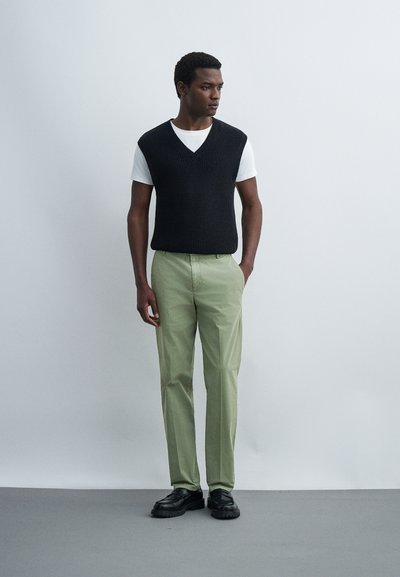 Άνδρας που φοράει μαύρο πλεκτό γιλέκο χωρίς μανίκια πάνω από λευκό T-shirt, ανοιχτοπράσινο παντελόνι και μαύρα παπούτσια, στέκεται με το ένα χέρι στην τσέπη μπροστά σε απλό τοίχο.