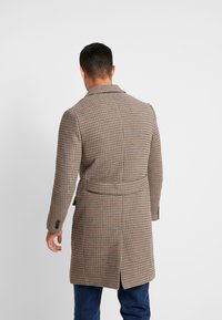 Hombre con un abrigo de cuadros marrón hasta las rodillas y respiraderos laterales, de espaldas contra un fondo blanco liso.