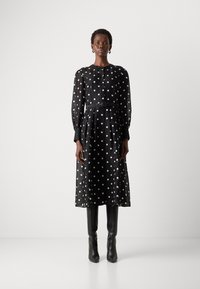 Abito nero a maniche lunghe con pois bianchi e cintura in vita. Tessuto trasparente e chiusura con bottoni. Abbinato a stivali neri al ginocchio.