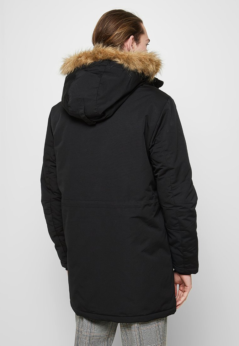 Parka negra con capucha forrada de piel, diseño acolchado, mangas largas y corte a media longitud, con una tela texturizada y sencilla.