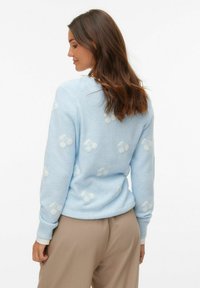 Jasnoniebieski sweter z białym wzorem wiśni, dekolt w łódkę, ściągacze przy rękawach i u dołu. Miękka faktura. Model nosi beżowe spodnie.