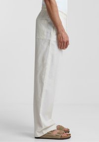 Byxor av linneblandning i ljus beige med avslappnad passform, med dragsko i midjan och en bakficka. Bärs med bruna sandaler.