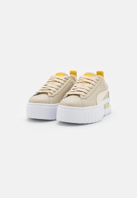 Sneakers platform beige con suole bianche spesse, lacci beige, linguette gialle sul tallone e cuciture decorative su uno sfondo bianco semplice.
