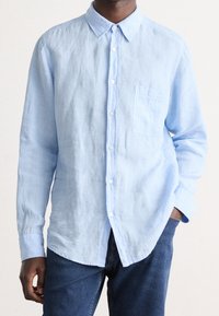 Camicia button-up in lino azzurro chiaro con colletto, taschino sul petto, e maniche lunghe; abbinata a jeans di jeans scuri.